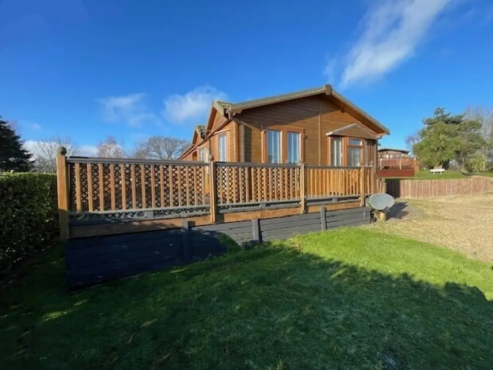 A wooden house with a deck, large windows, a satellite dish, and a grassy yard under a partly cloudy blue sky.