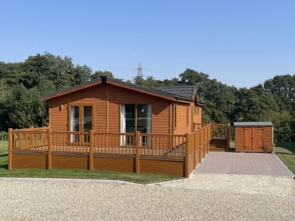 A wooden cabin with a deck and railing, set on a grassy lawn with a paved area and a small garden shed, surrounded by trees under a clear blue sky.