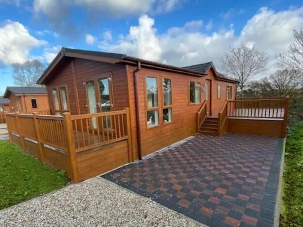 A wooden house with large windows, a spacious deck, and a checkered driveway under a partly cloudy sky.