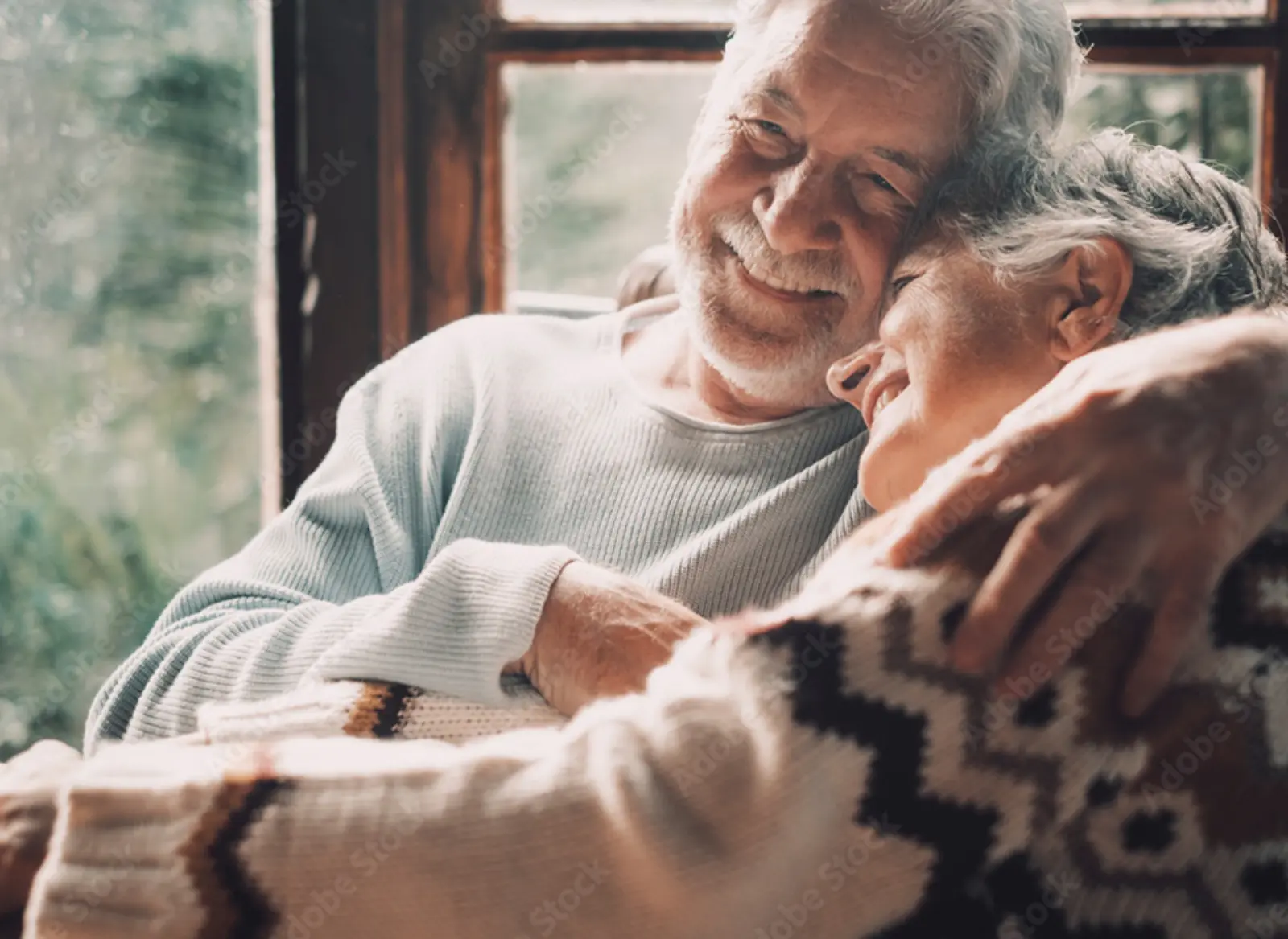 An elderly couple sharing a warm, joyful embrace near a window, smiling and looking happy together in a cozy setting.