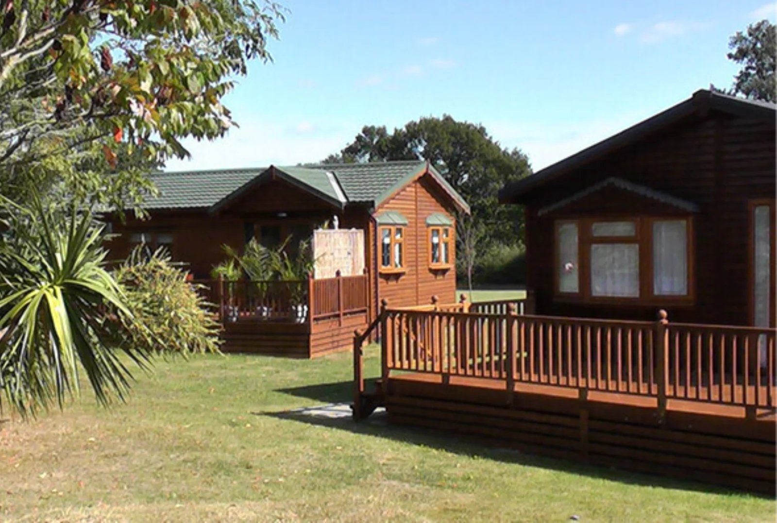Two wooden cabins with decks are surrounded by green grass and trees under a blue sky.