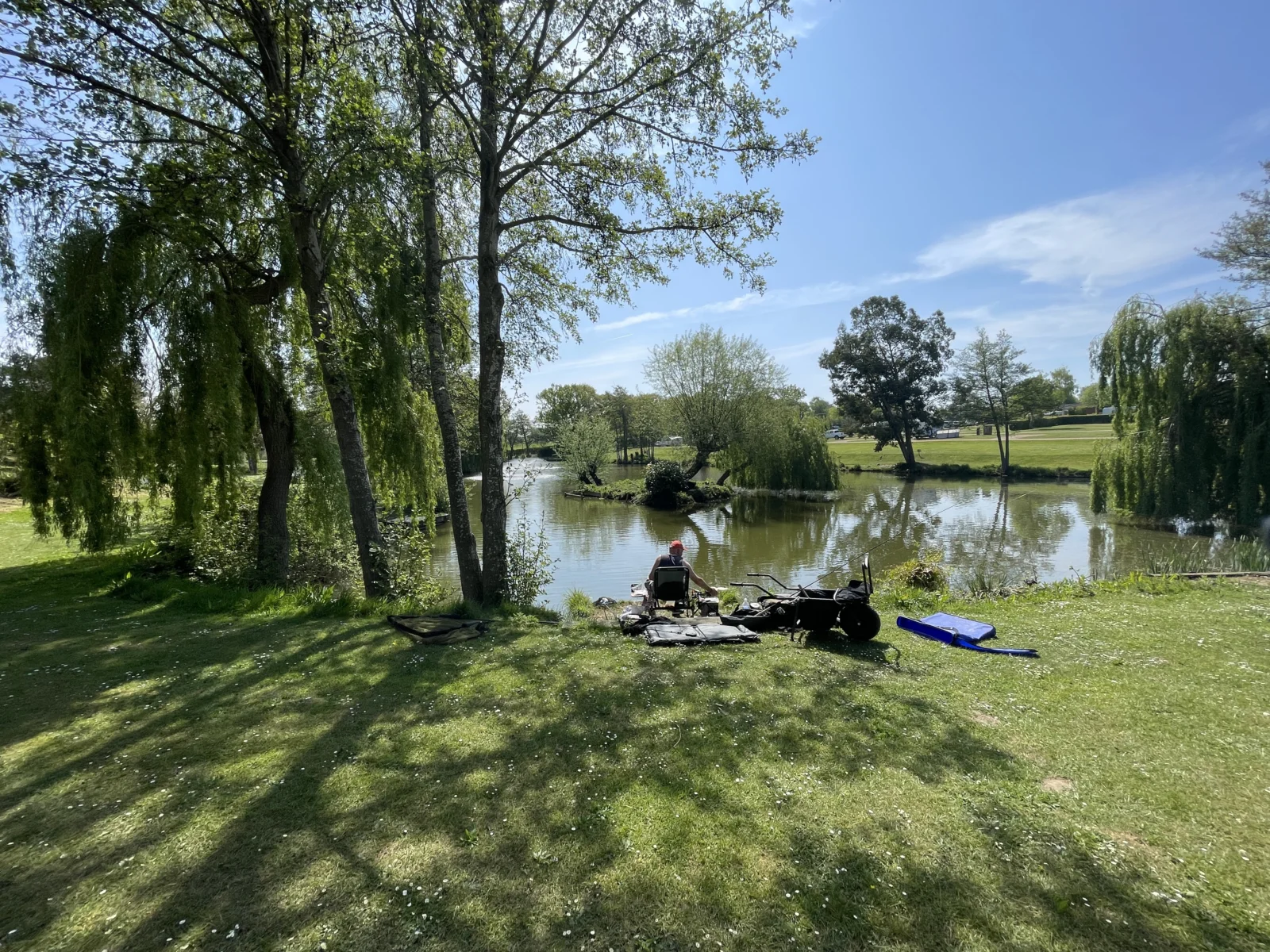 Person sitting by a pond surrounded by trees, with fishing gear and a blue float on the grassy bank under a clear blue sky.