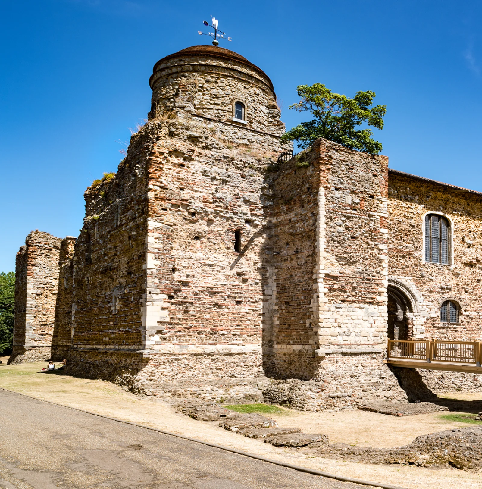 Ancient stone castle tower under a clear blue sky with a tree beside it, part of a larger fortress structure with a wooden bridge in front.