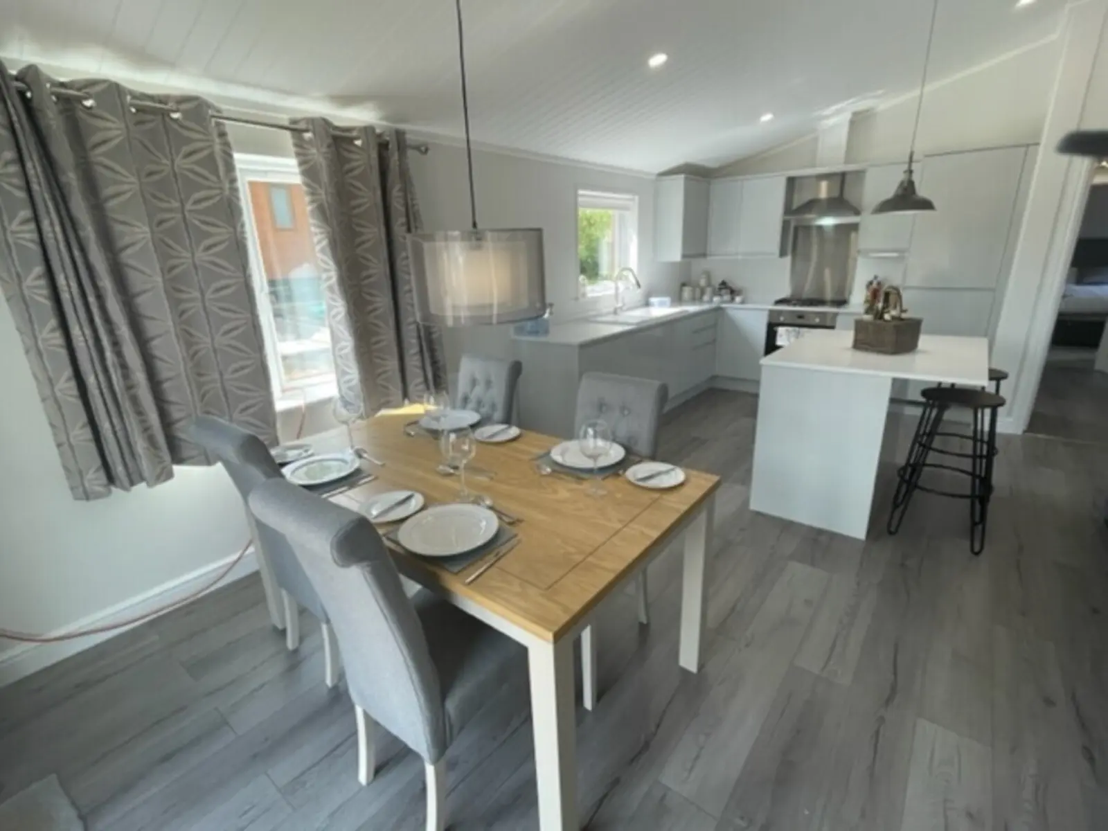 Open-concept kitchen and dining area with a wooden table set for four, gray upholstered chairs, white cabinetry, and pendant lighting.