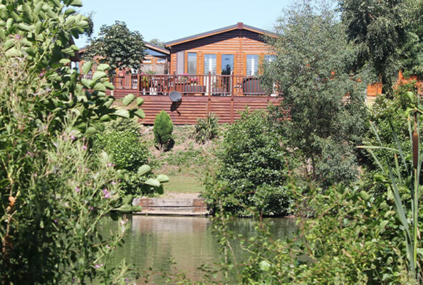 A wooden house with a deck, surrounded by lush greenery and trees, overlooking a small pond in a peaceful garden setting.