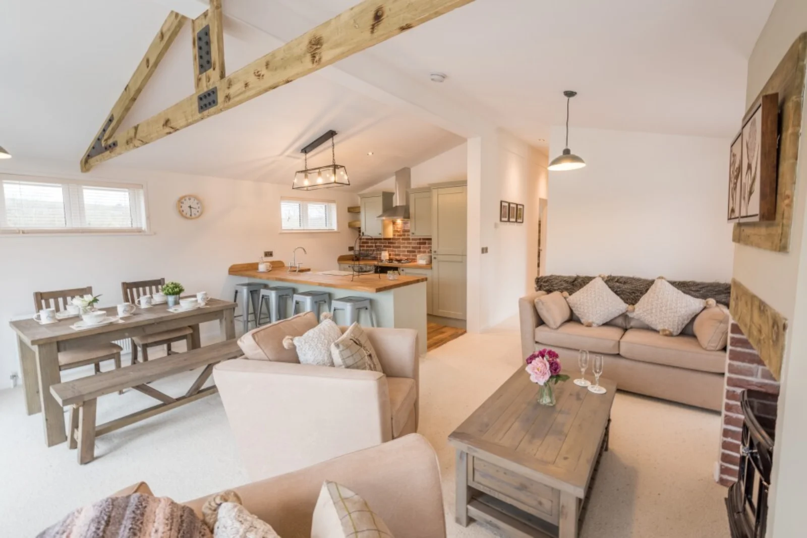 Open-plan living and dining area with a kitchen, featuring beige sofas, wooden furniture, exposed brick, and decorative pillows, bright and cozy ambiance.