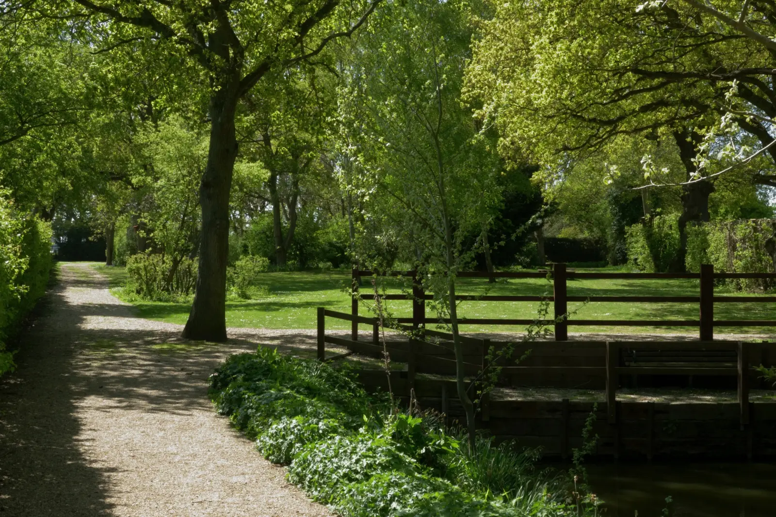 A peaceful park scene with a gravel pathway, lush green trees, a small wooden bridge over a waterway, and sunlight filtering through the foliage.