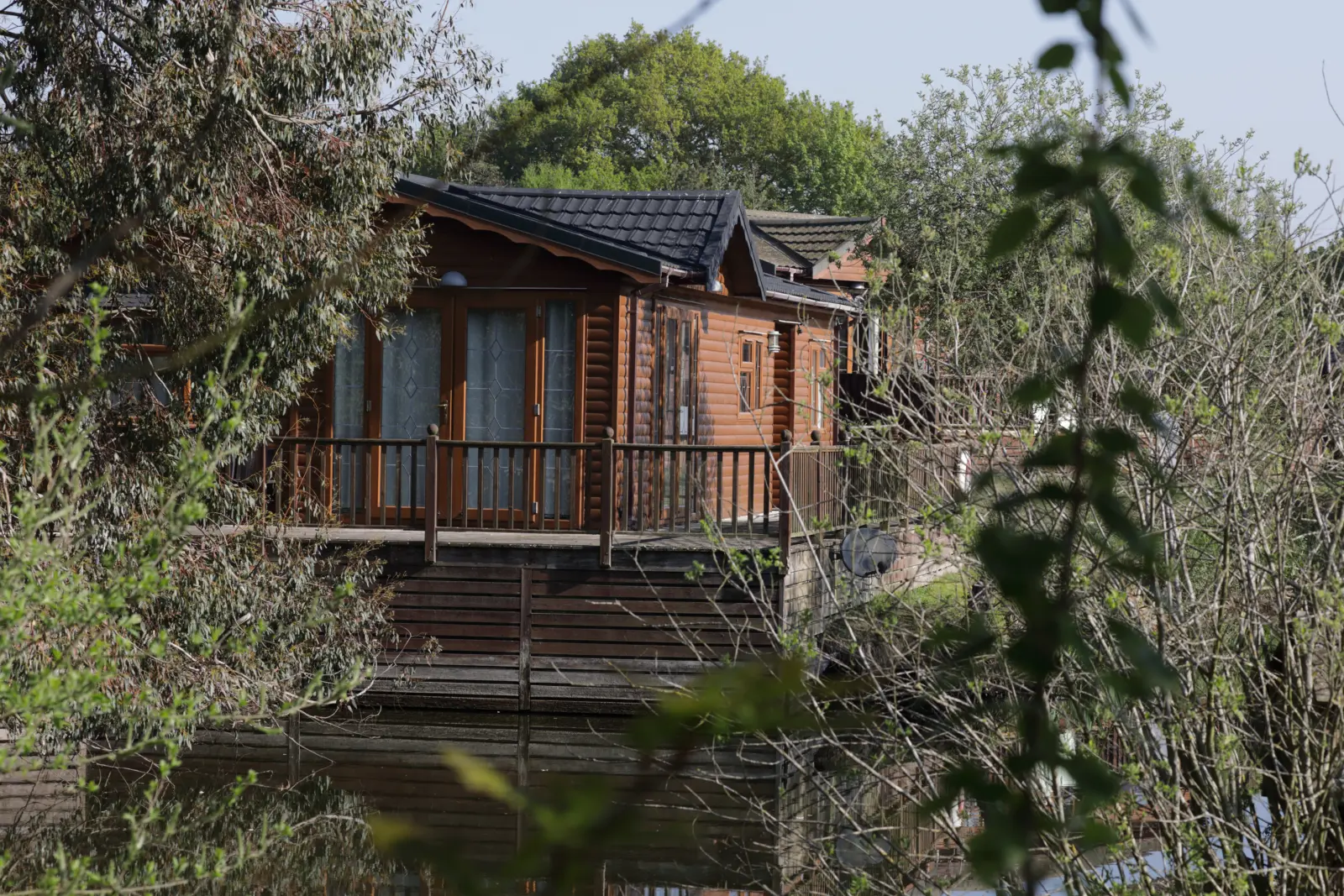 A wooden house on stilts surrounded by trees and foliage, with a balcony and reflective water below, appears to be in a wooded, lakeside setting.