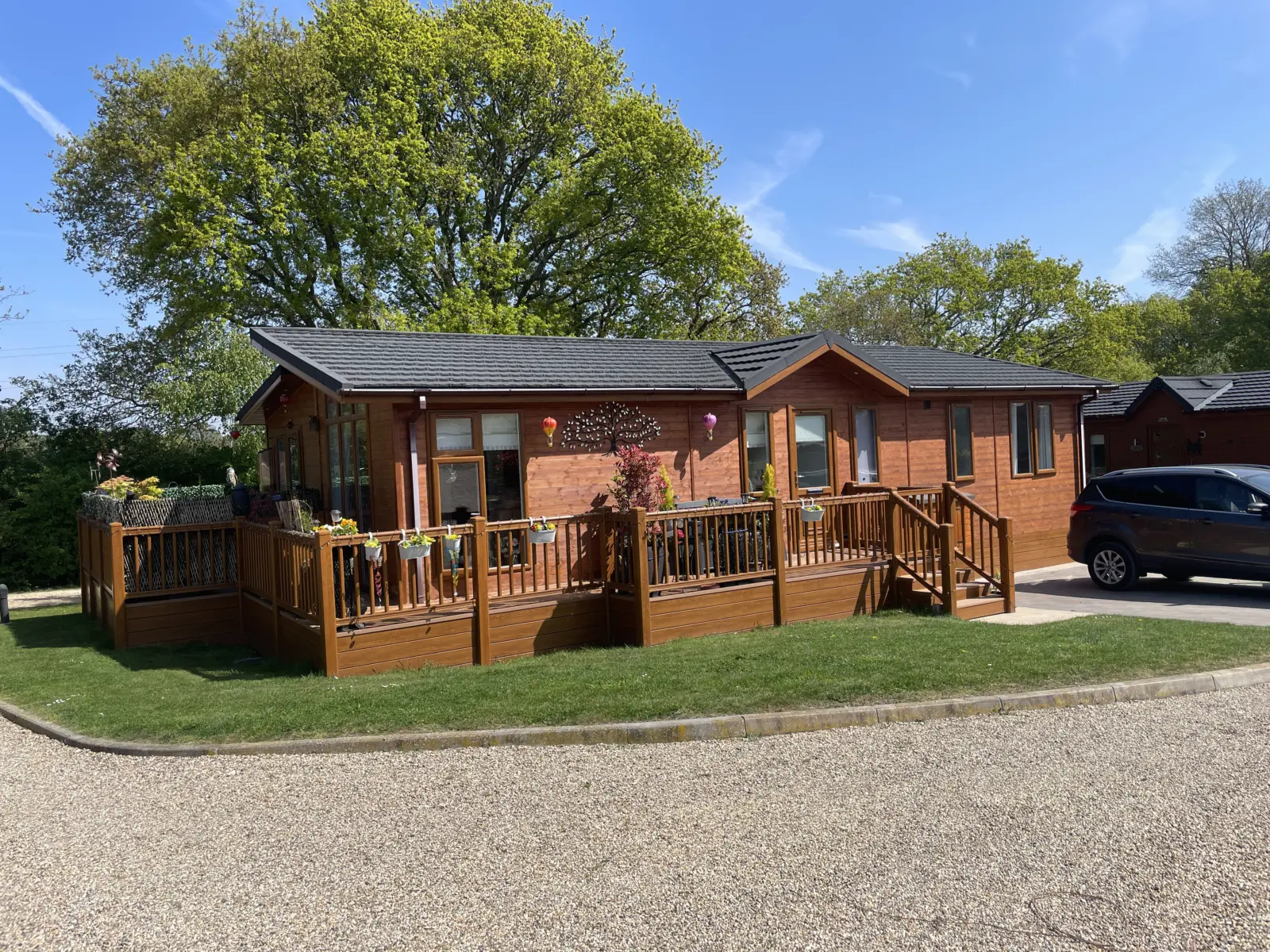 A wooden house with a large deck, decorated with hanging planters and ornaments, surrounded by green trees and a parking area with a black car.