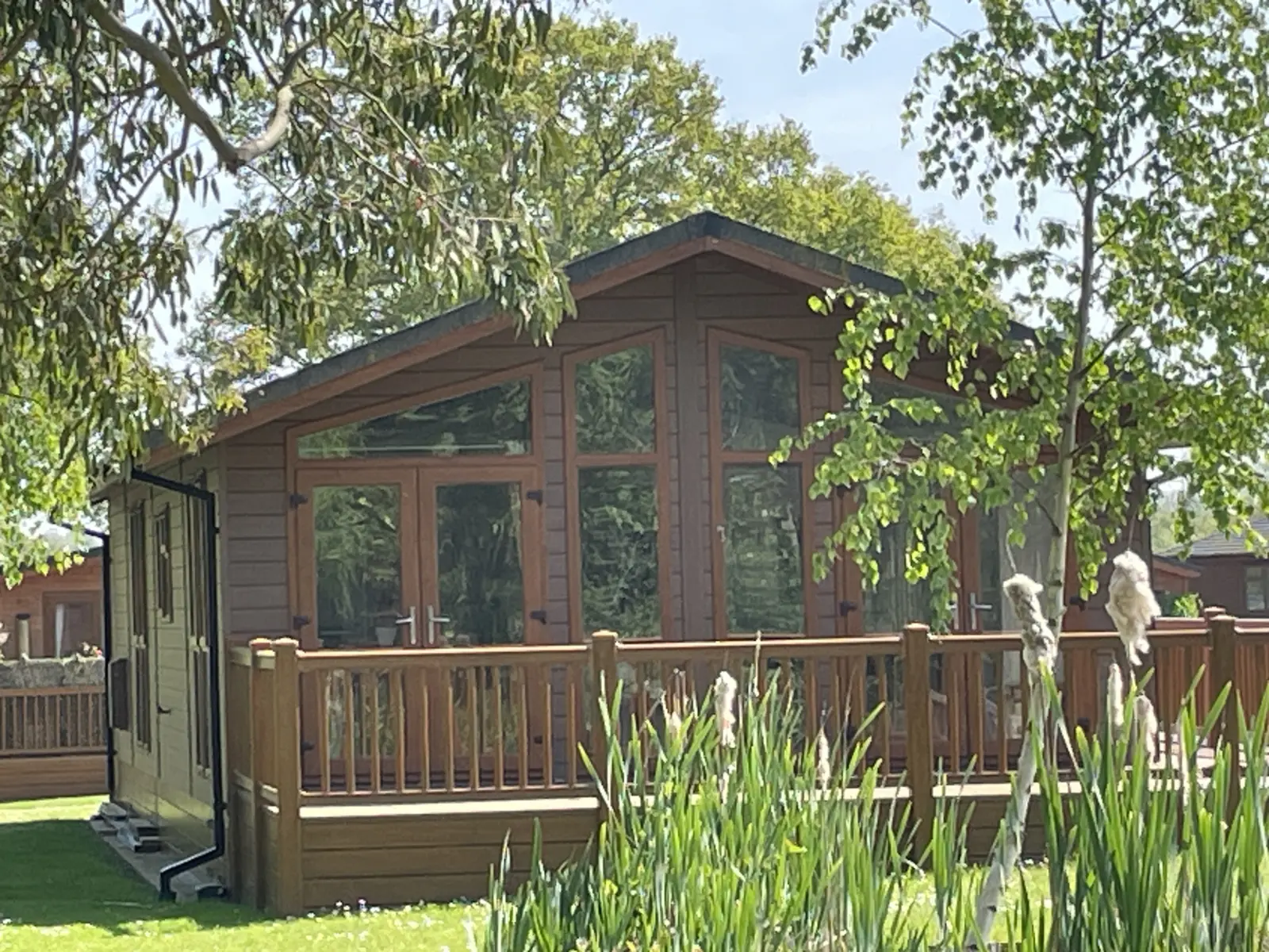 A cozy wooden house with large glass windows, surrounded by a wooden deck, lush green trees, and tall grass in a sunny backyard.