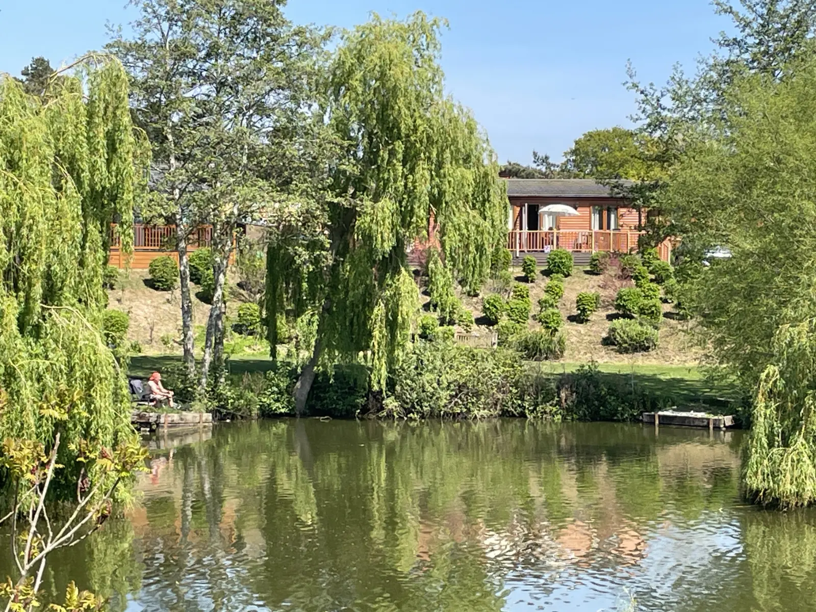 A person sitting on a dock by a calm pond surrounded by lush green trees and a hillside with a wooden house and garden in the background.