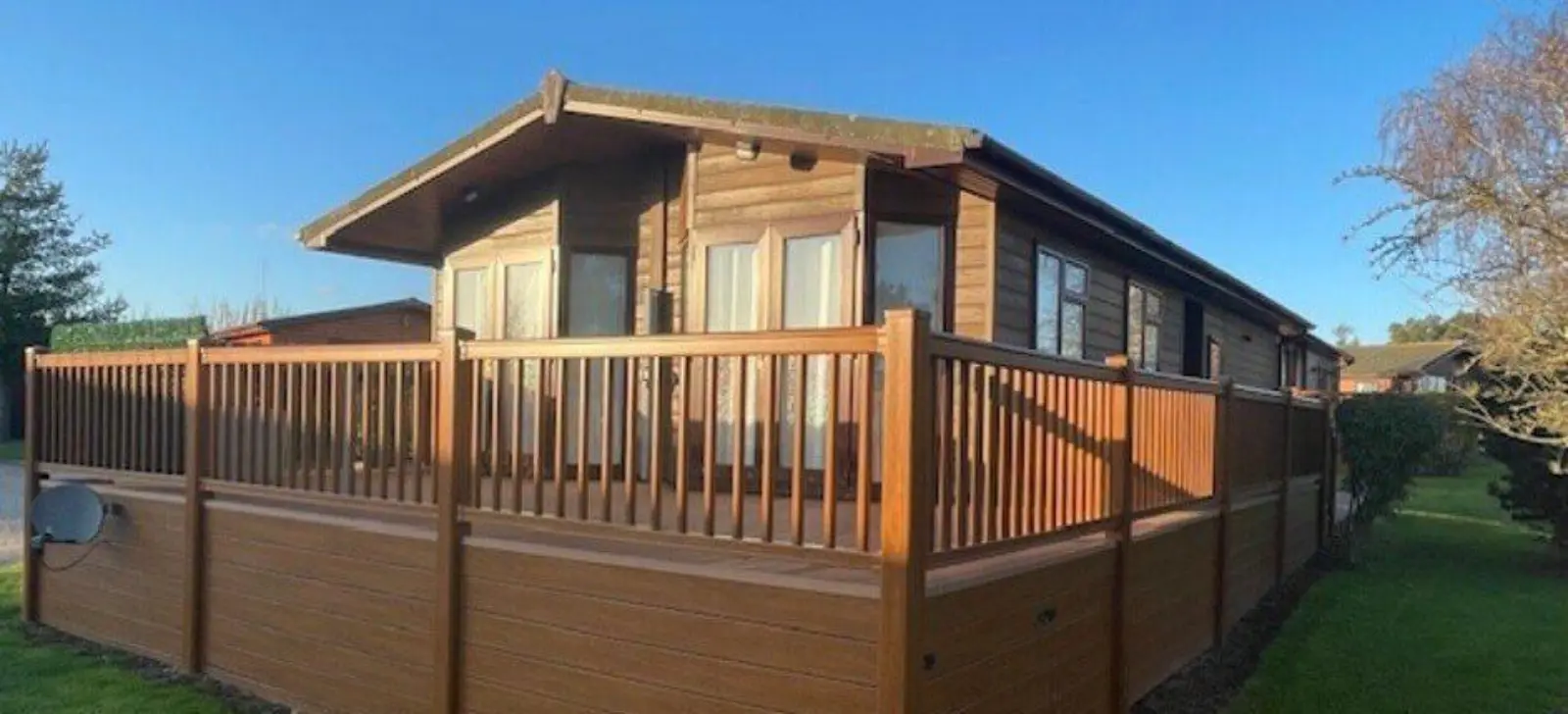 A wooden house with large windows and a spacious balcony, surrounded by a tall wooden fence and a well-maintained lawn, under a clear blue sky.