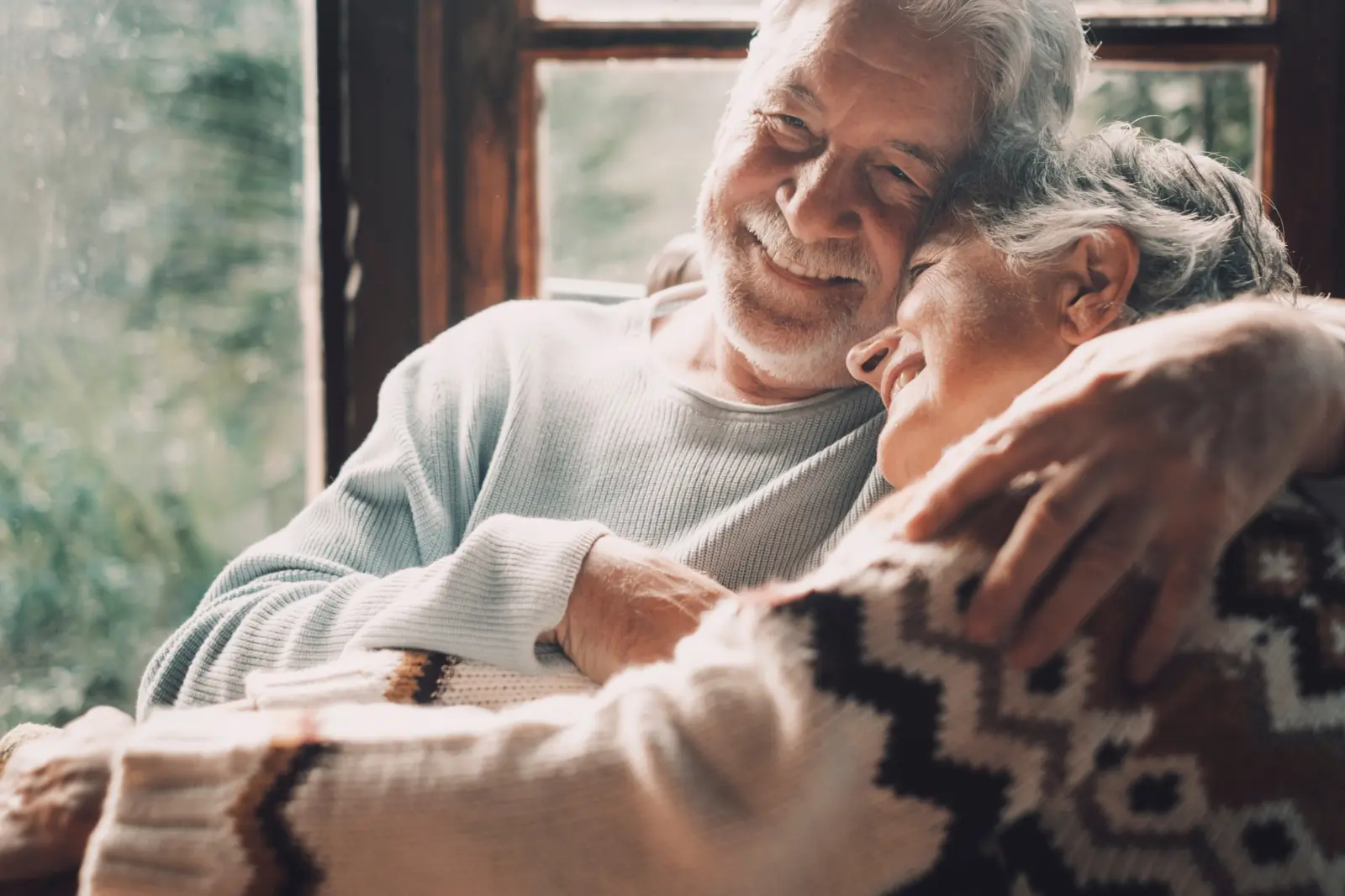 An elderly couple shares a warm embrace and smiles by a window, creating a cozy, joyful moment of companionship and love indoors.