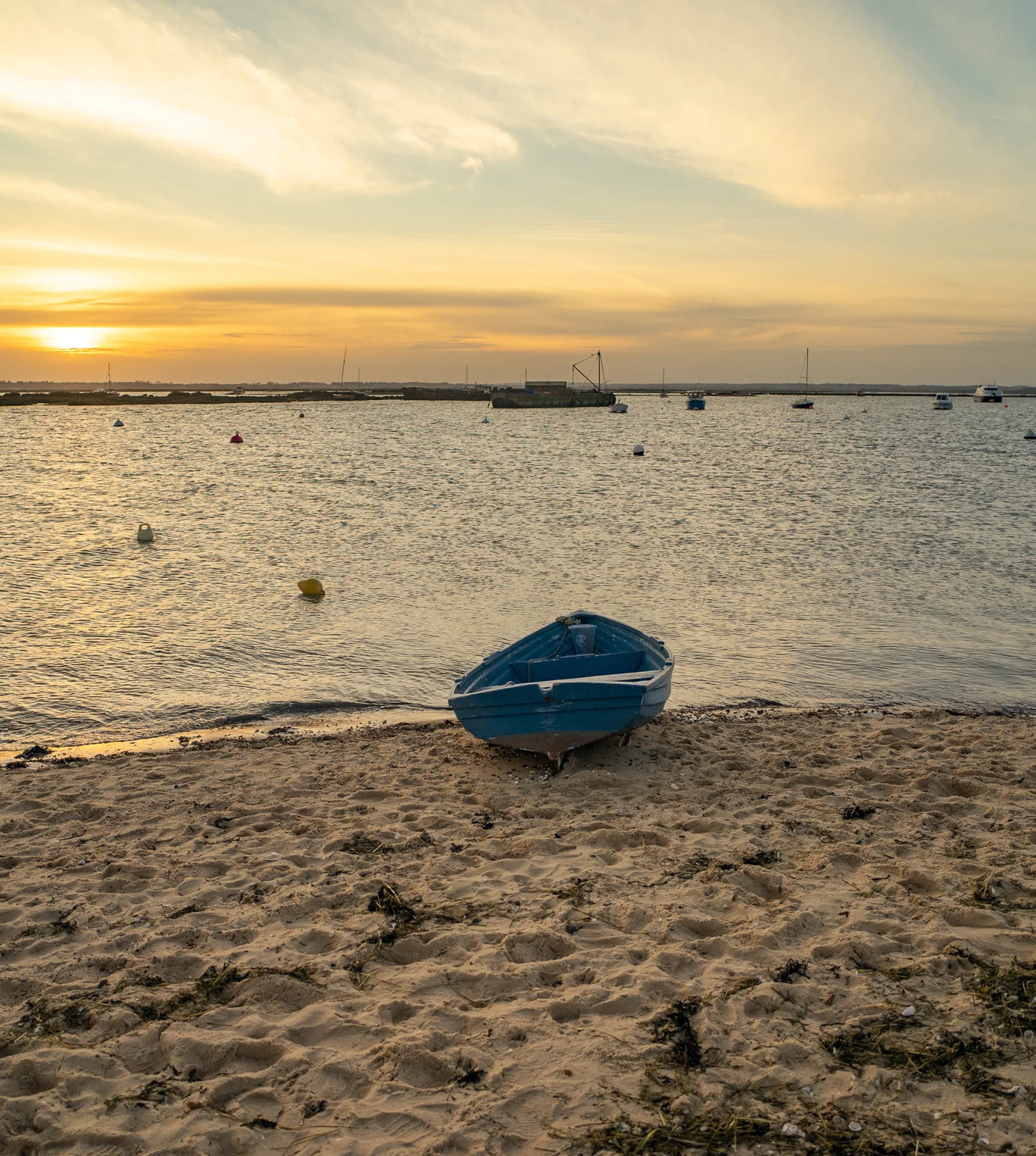 A small blue boat rests on a sandy beach at sunset, with calm water and distant boats and a cloudy sky in the background.