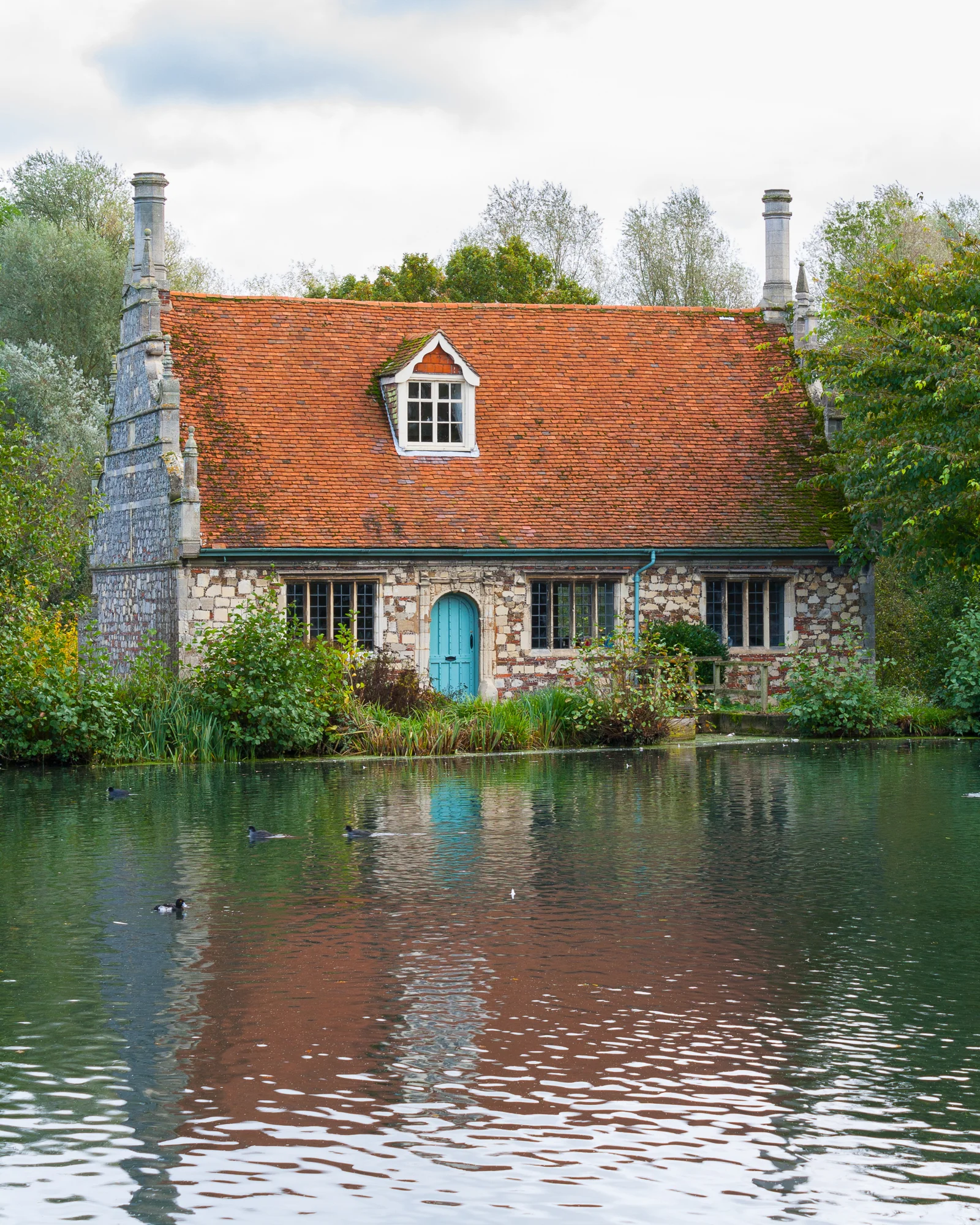 A stone cottage with a red tiled roof, blue door, and multiple windows sits beside a pond with ducks, surrounded by lush greenery and trees.