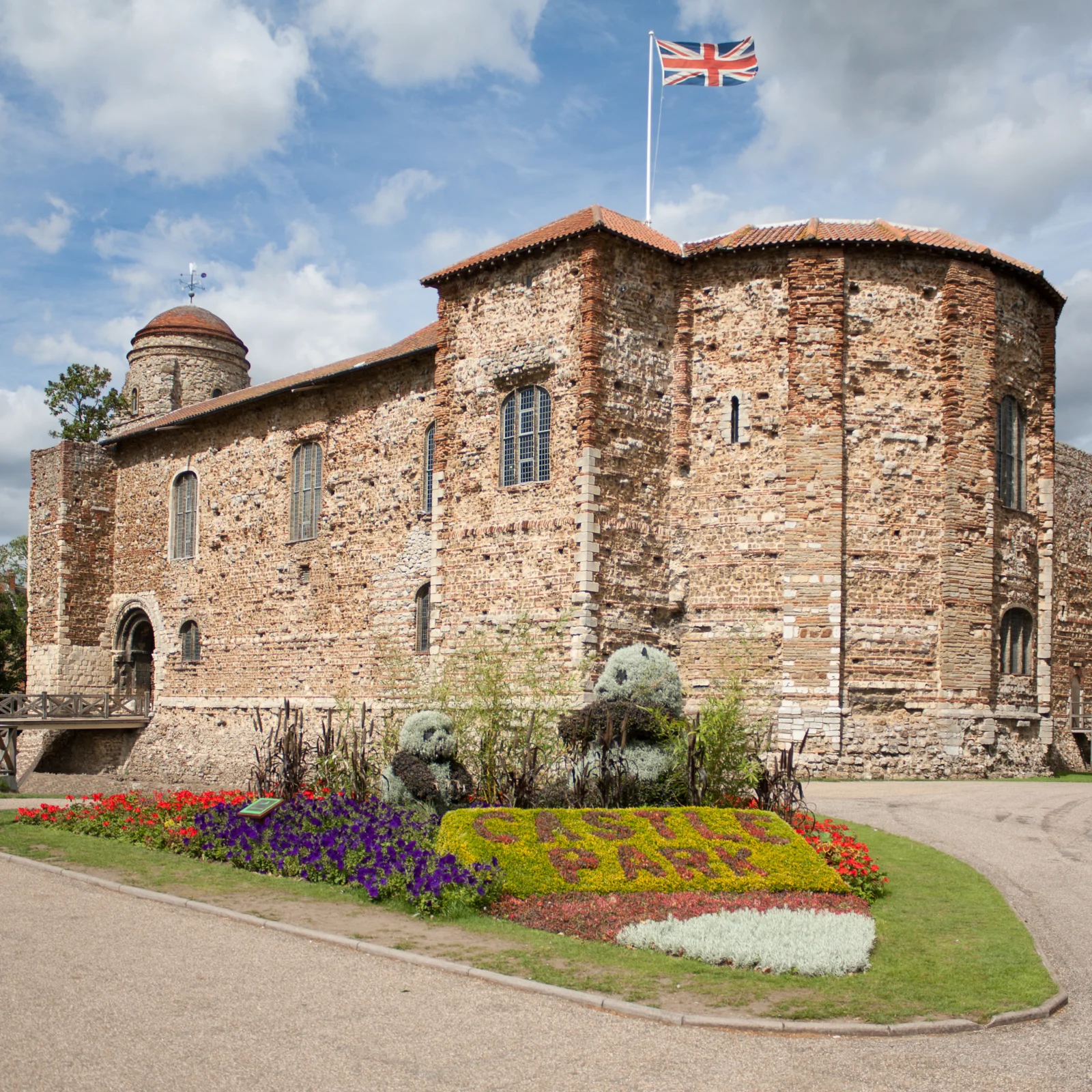A historic stone castle with small windows and a Union Jack flag on top, surrounded by colorful flowers and a clear sky with some clouds.