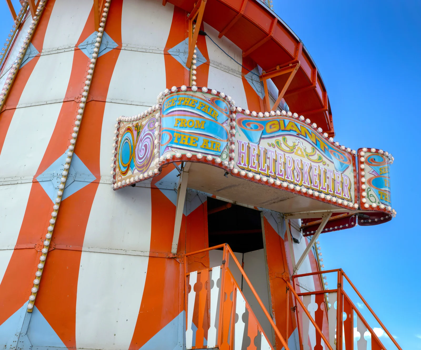 A colorful fairground ride with a sign reading "Giant Helterskelter" and "See the fair from the air," set against a bright blue sky.