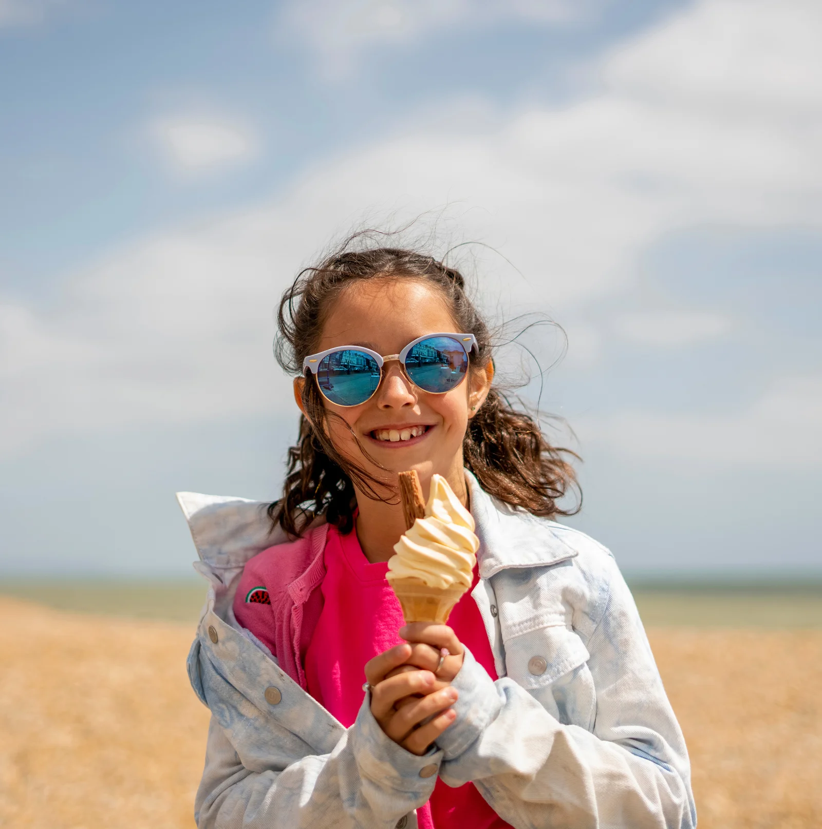 Smiling girl wearing sunglasses, holding a vanilla ice cream cone with a biscuit on top, on a beach with cloudy sky in the background.
