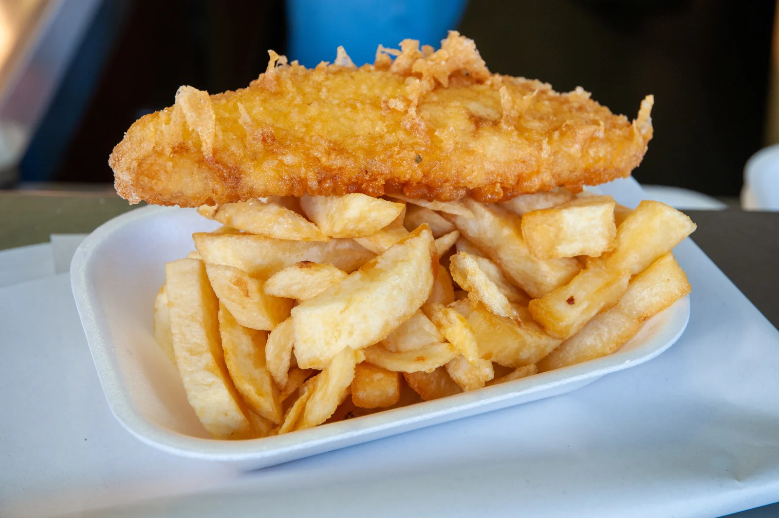 A styrofoam container filled with crispy, golden French fries topped with a large, crispy fried fish fillet on a table.
