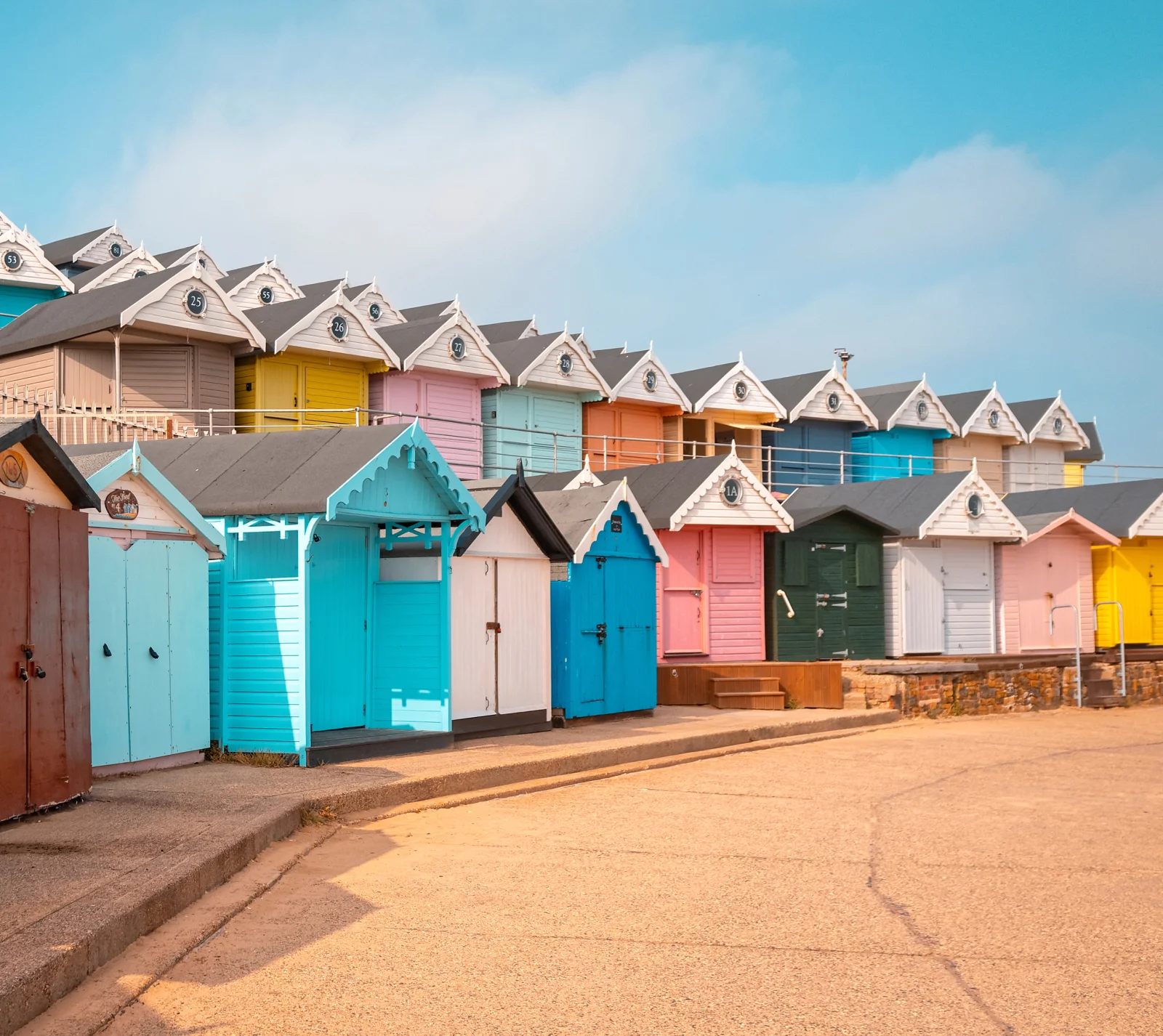Colorful beach huts lined up along a sandy pathway, with a bright blue sky in the background, creating a cheerful seaside scene.