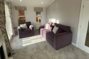 A cozy living room with two purple sofas, light-colored walls, floral curtains, and sunlight streaming through windows, creating a warm and inviting atmosphere.