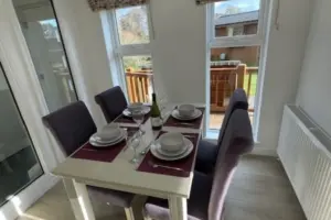 Dining table set for four with plates, glasses, wine bottle, and cutlery in a bright room with large windows and a sliding door leading to an outdoor deck.