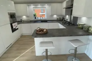 Modern white kitchen with a dark countertop island, two white bar stools, and a window with a small plant in the background.