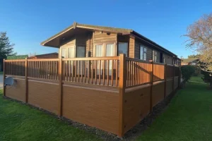 A wooden house with a large deck surrounded by a wooden railing, set in a grassy yard under a clear blue sky.