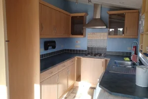 Small kitchen with wooden cabinets, black countertops, a stovetop, stainless steel extractor, and window above the sink, with various appliances and outlets.