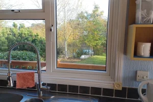View through a kitchen window showing trees, shrubs, and a grassy yard outside with a small pond or water feature in the distance.