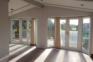 Bright living room with large windows and glass doors leading to a balcony, allowing sunlight to cast shadows on the carpeted floor.