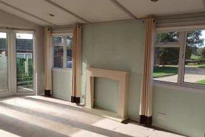 Empty room with large windows and a glass door, sunlight casting shadows on the carpet, fireplace mantle, and views of a garden outside.