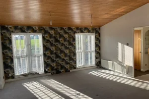 Empty living room with three large windows, patterned accent wall, wood ceiling, and sunlight casting shadows on the beige carpet.