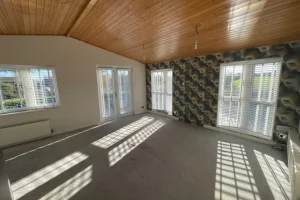 Bright, spacious living room with large windows and wooden ceiling, sunlight casting shadows on the carpeted floor, patterned accent wall on the right.