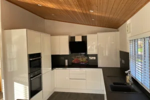Modern kitchen with white cabinets, black countertops, built-in appliances, a black range hood, and a window with blinds allowing natural light.