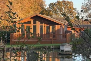 A wooden house with large windows and a deck overlooks a calm pond, surrounded by trees with autumn-colored leaves, reflected in the water.