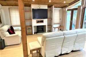 Living room with cream-colored sofas, wooden accents, a television above a fireplace, and large windows with curtains letting in natural light.