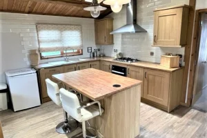 A cozy kitchen with wooden cabinets, a central island with seating, a window with a patterned blind, and a ceiling with wooden paneling and hanging lights.