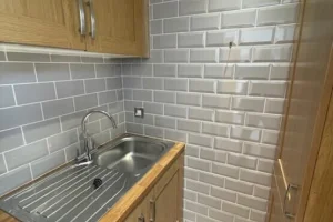 Small kitchen corner with wooden cabinets, a stainless steel sink, gray brick backsplash, and light wooden flooring.