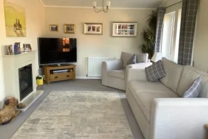 Living room with a beige sofa, armchair, TV on a wooden stand, fireplace, framed pictures on the wall, curtains, and a large window with blinds.