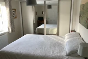 A bedroom with a bed covered in white bedding, a bedside lamp, a window with patterned curtains, and a mirrored wardrobe reflecting the room.