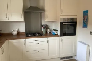 Kitchen with cream cabinets, wooden countertop, gas stove, built-in oven, and various kitchen items on the counter. A radiator is on the right wall.