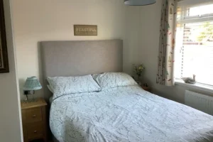 A neatly made bed with a beige headboard, white bedspread, and pillows in a room with a window and patterned curtains, a bedside table with a lamp, and a small vase of flowers.