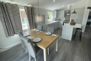 Open-concept kitchen and dining area with a wooden table set for four, gray upholstered chairs, white cabinetry, and pendant lighting.