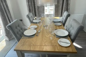 A wooden dining table set for six with white plates, silverware, and empty wine glasses, surrounded by grey upholstered chairs in a bright room.