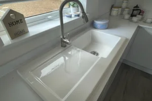 White kitchen sink with a faucet, set in a countertop near a window with kitchen jars and a decorative "HOME" sign on the windowsill.
