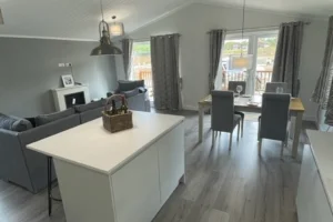 Bright living and dining area with grey chairs, curtains, and a large window showing an outdoor deck. Light-colored flooring and furniture.