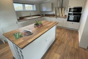 Modern kitchen with white cabinets, a wooden island, stainless steel appliances, and wood flooring, illuminated by natural light from windows.