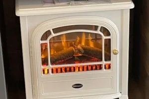White electric fireplace with orange flames and logs, placed against a dark wooden floor and wall, with a small top surface holding papers and magazines.