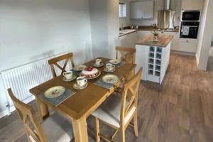 Dining area with a wooden table set for five, in an open-plan kitchen with white cabinets, wooden floors, and a skylight overhead.